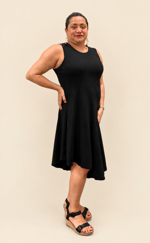 Full-length portrait of LM D’Lishes in a sleeveless black dress and black wedge sandals, posing confidently with one hand on her hip against a solid cream background.